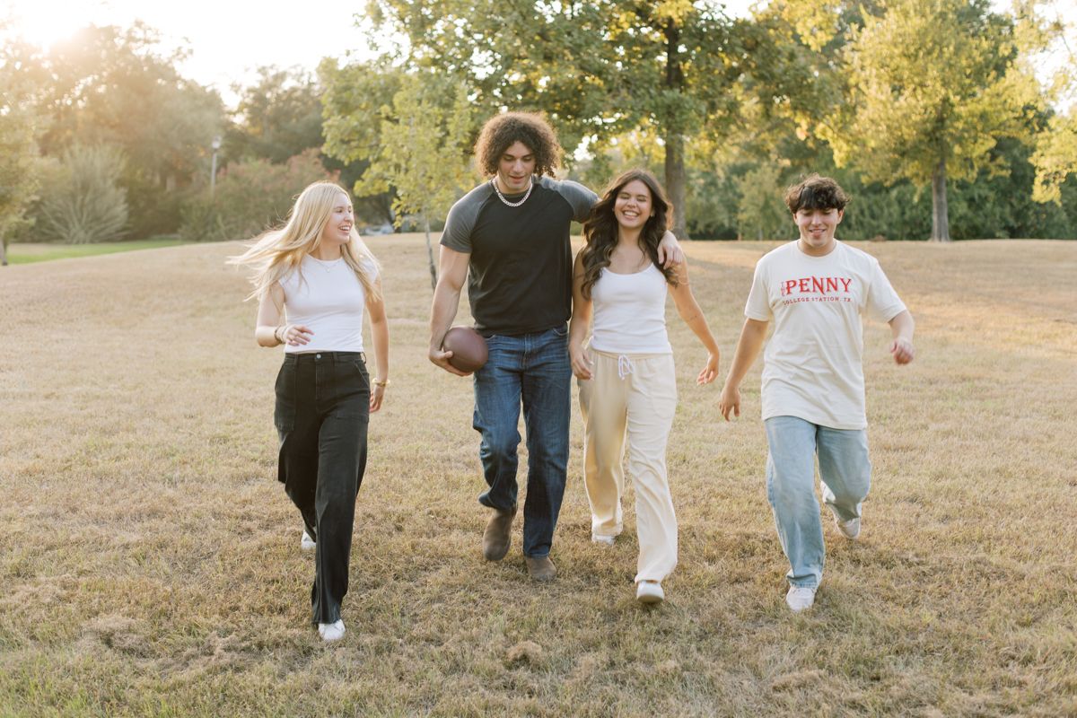 Four young friends walk together laughing in a sunny park with green trees in the background.