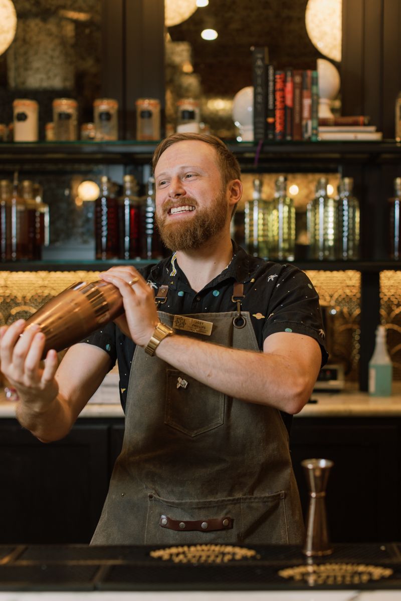Smiling bearded bartender in apron shaking a copper cocktail shaker behind a bar.