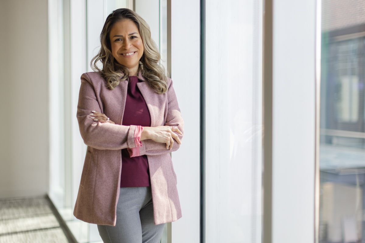 Confident businesswoman in pink blazer smiling with arms crossed by bright office windows.