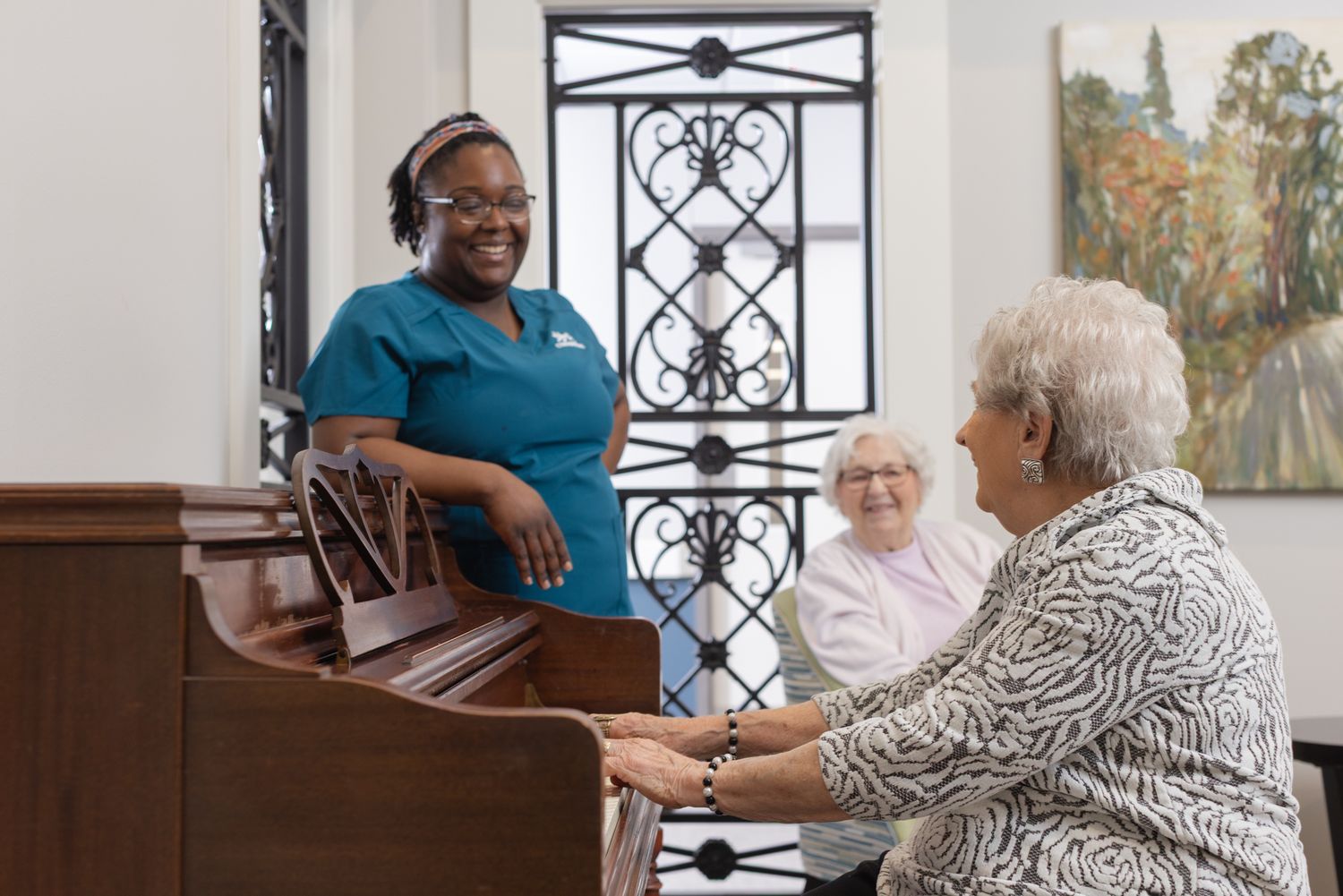 A caregiver smiles as an elderly woman plays piano while another resident watches nearby.