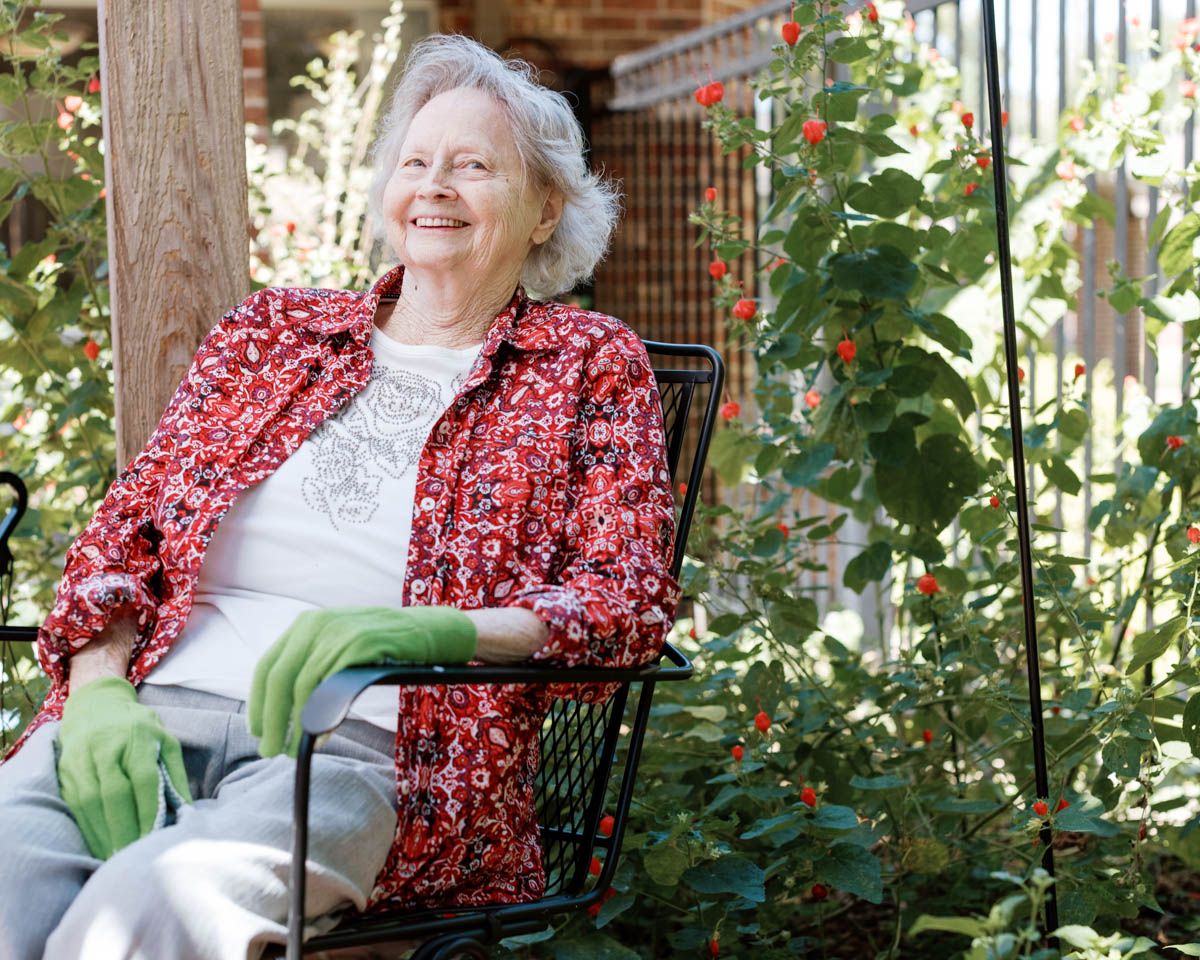 Happy senior woman sitting in garden chair surrounded by lush green plants and red flowers.