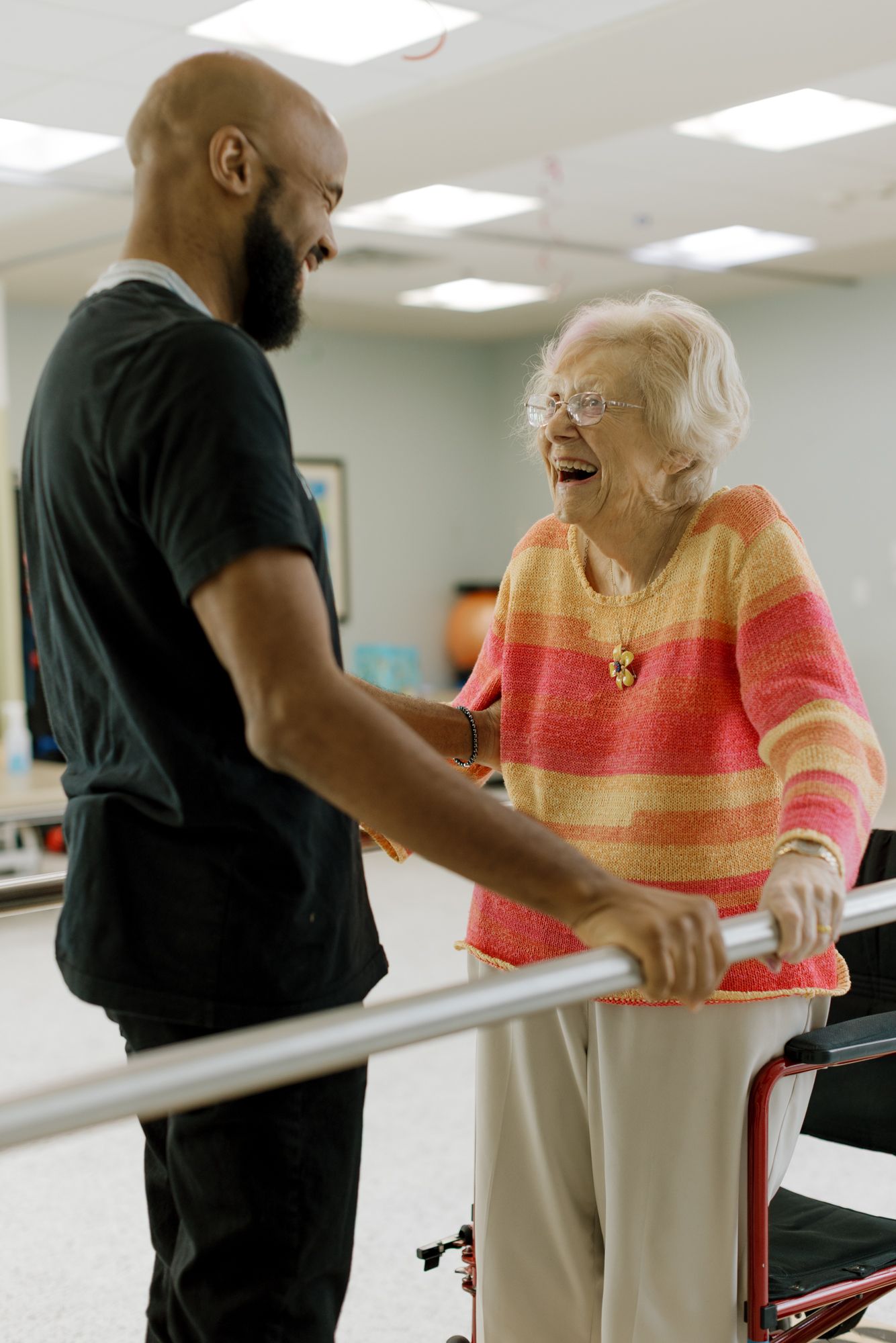 Elderly woman laughs with a bald bearded therapist while using parallel bars in a rehabilitation facility.