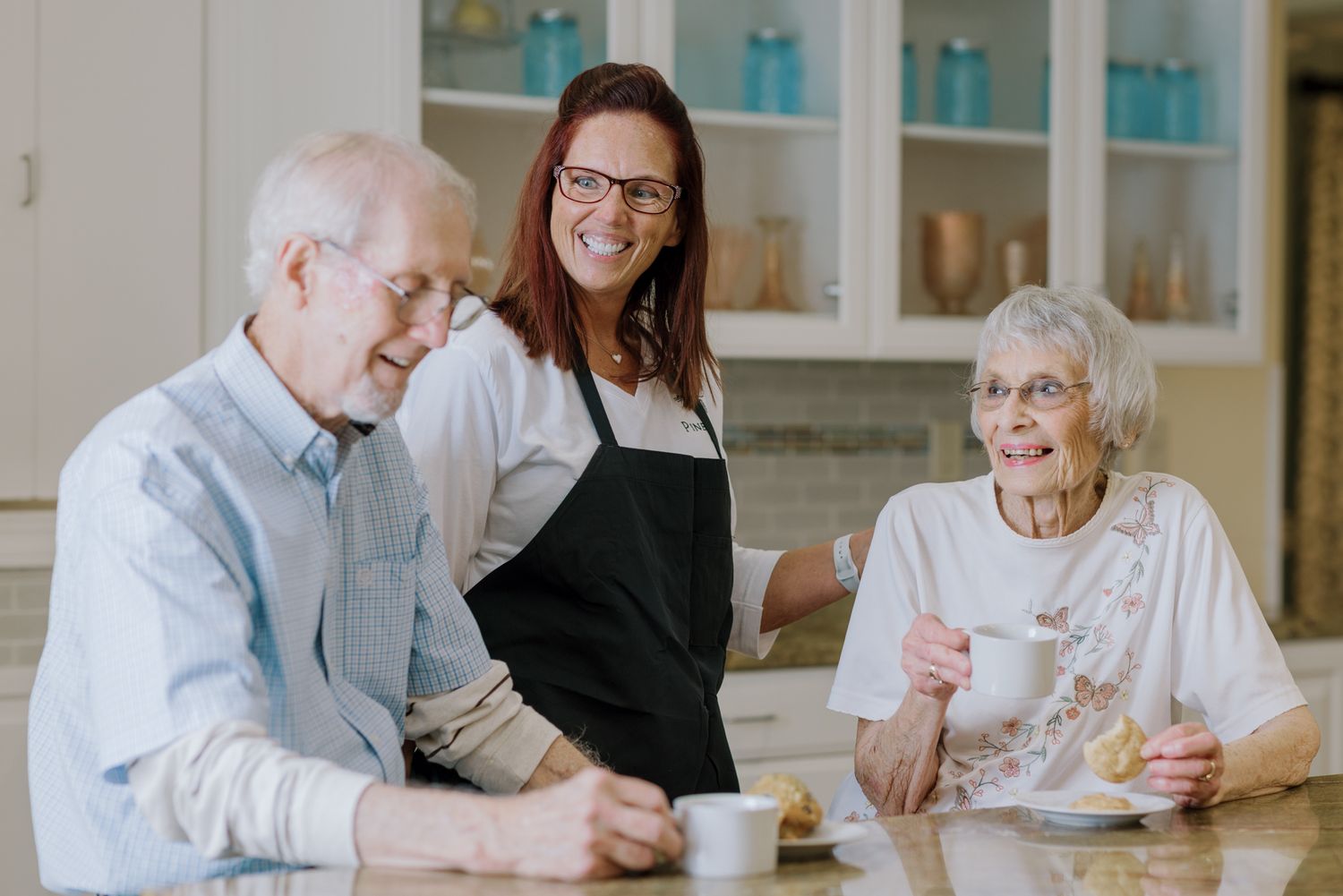 Caregiver in black apron sharing a joyful moment with elderly couple over coffee.