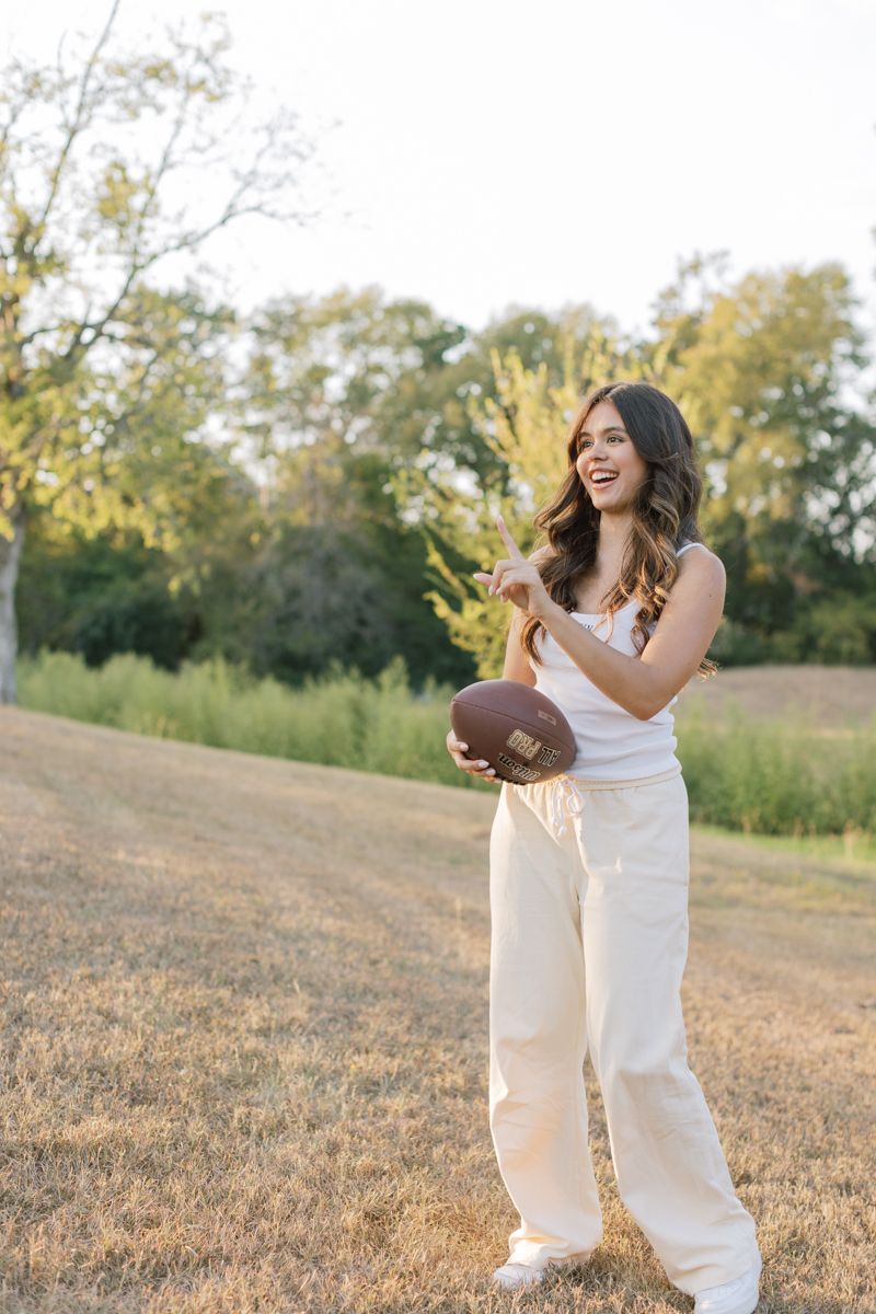 Smiling young woman holding a football in a sunny outdoor park setting.