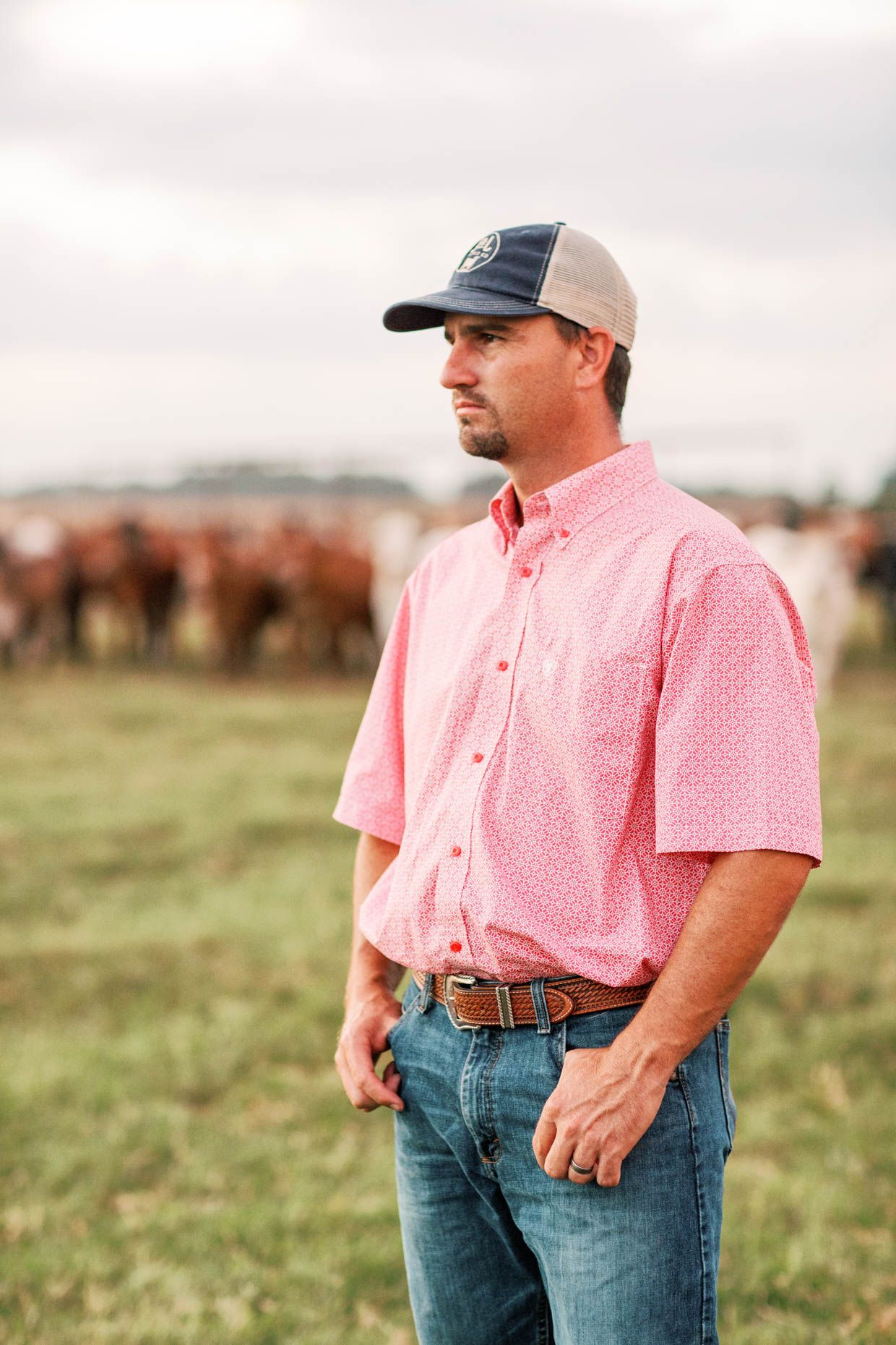 A rancher in a pink shirt and trucker hat stands in a field with cattle grazing in the background.