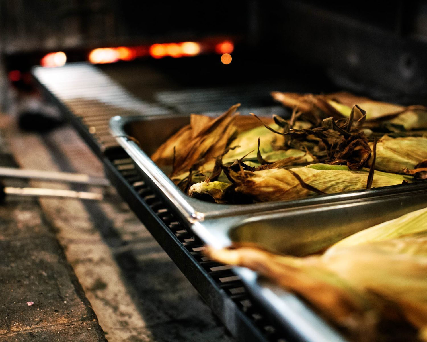 Corn tamales cooking on a grill tray beside a glowing wood-fired oven.