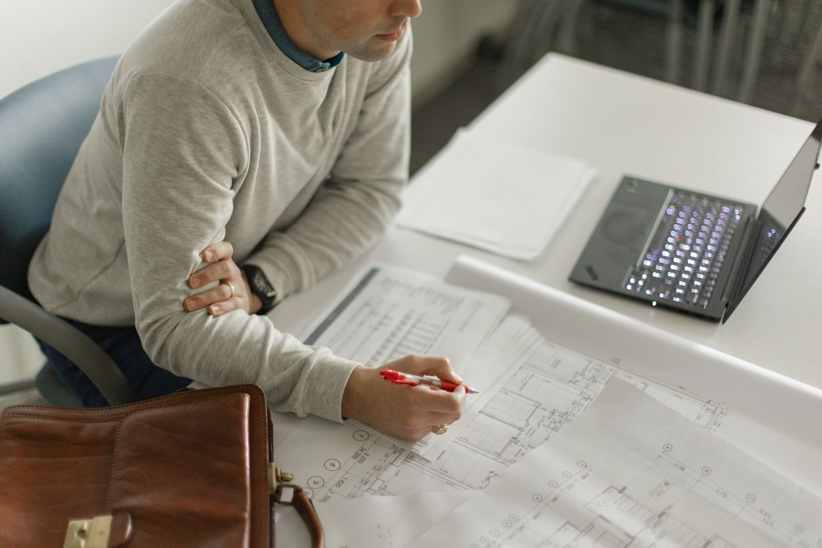 Architect reviewing blueprints at desk with laptop, holding red pen, brown leather bag nearby.