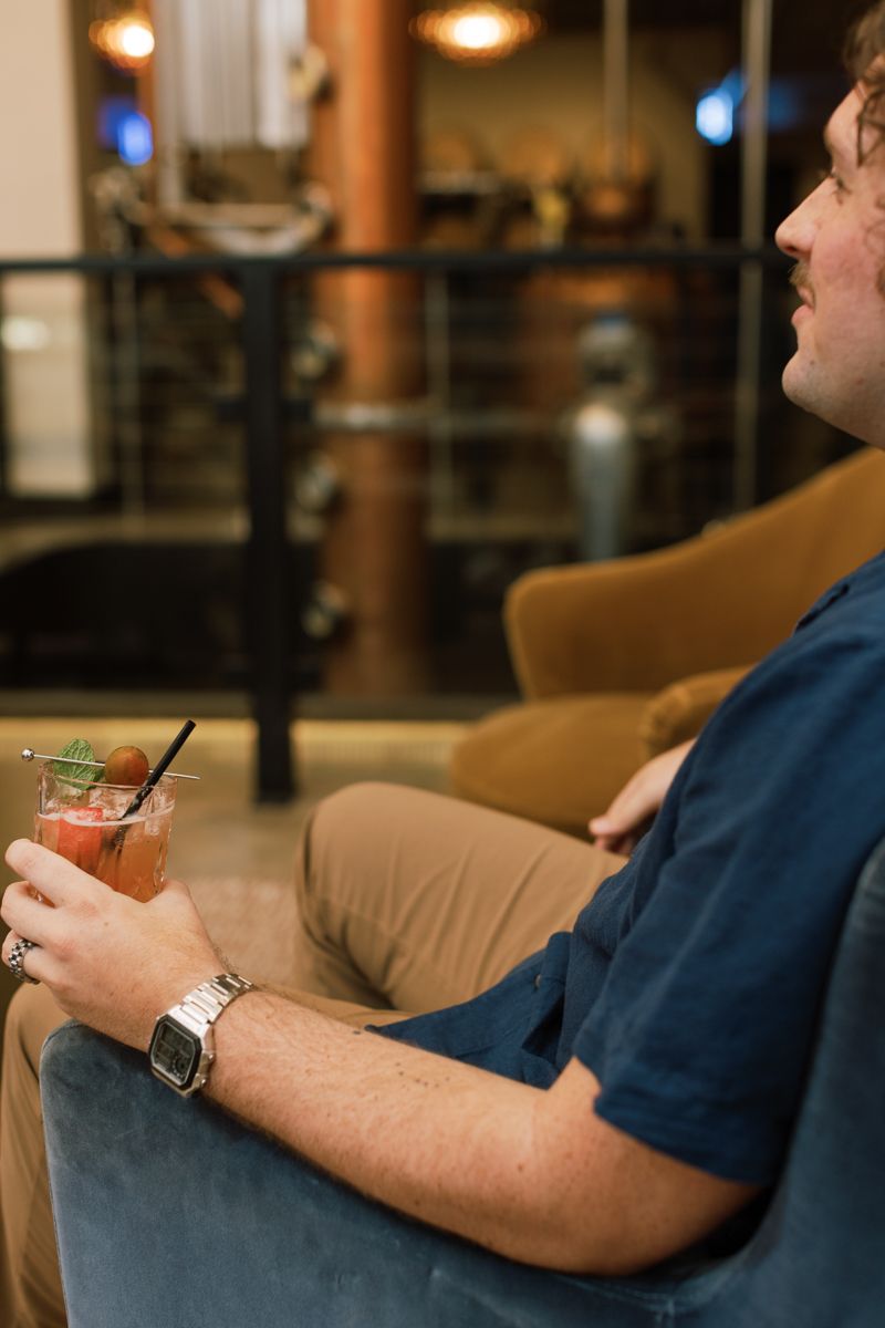 Man in blue shirt holding a pink cocktail with garnish, relaxing in a velvet chair at an upscale bar lounge.
