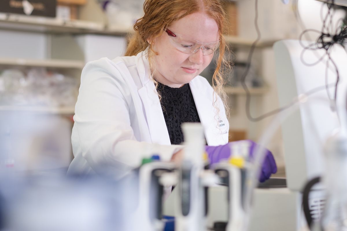 Female scientist with red hair in white lab coat working with microscope in research laboratory.