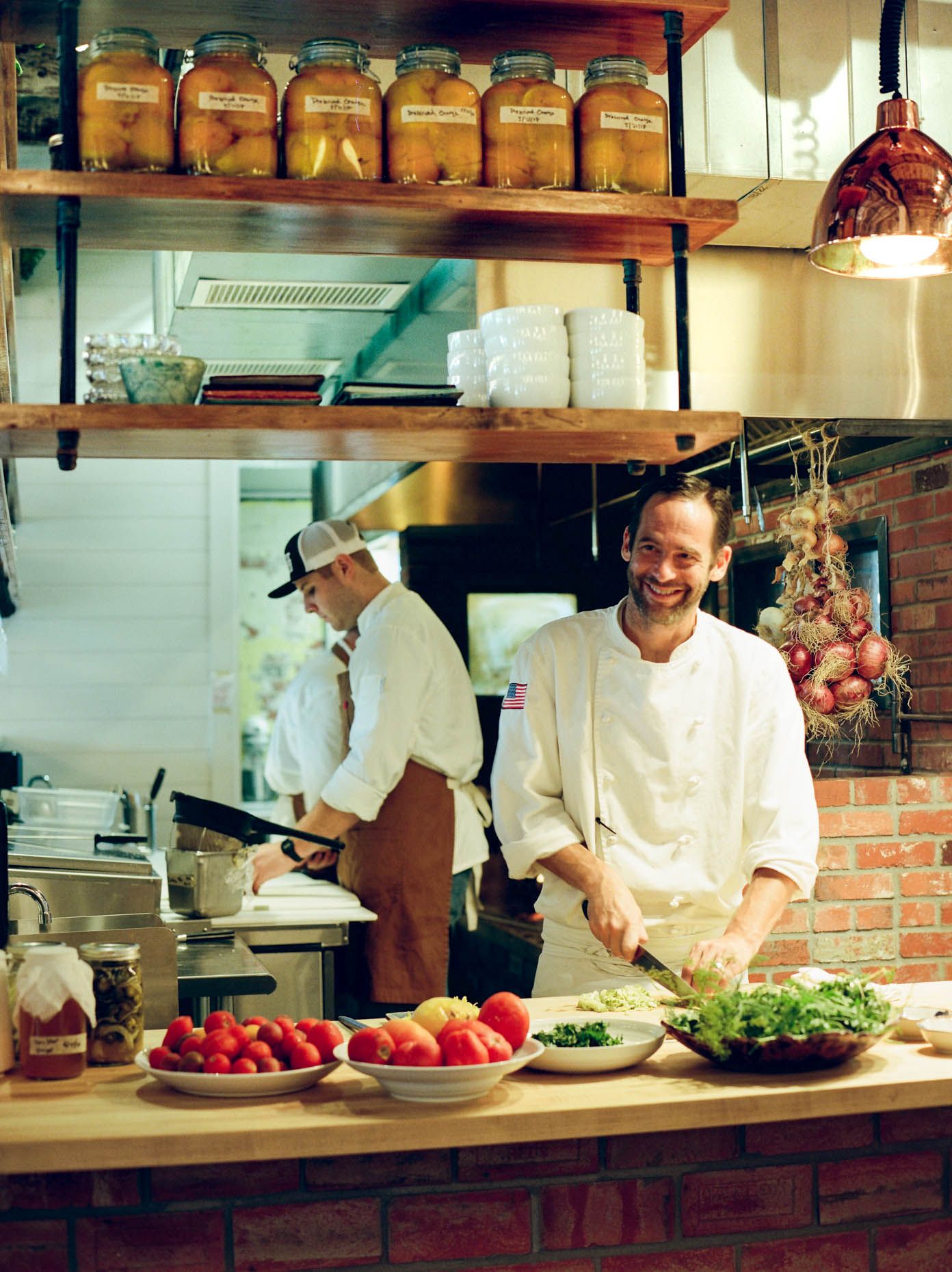 Smiling chef preparing fresh vegetables in a rustic kitchen with shelves of preserved foods and hanging onions.