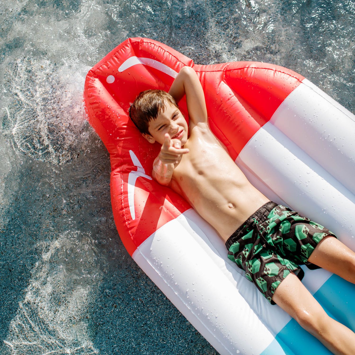 Boy relaxing on a red and white inflatable pool float in sparkling blue water, pointing at camera.
