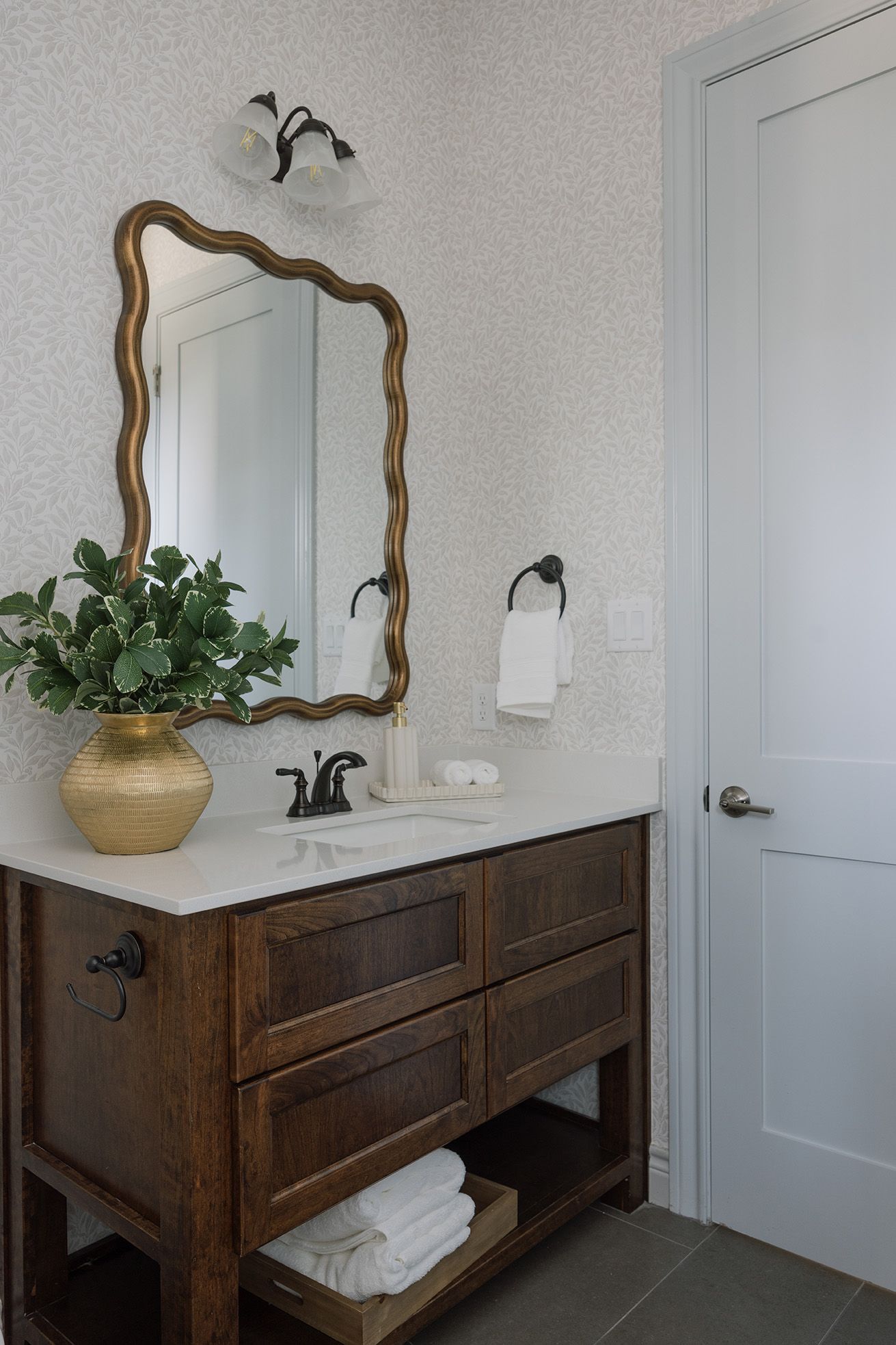 Dark wood bathroom vanity with wavy gold mirror, textured wallpaper, and brass vase with greenery.