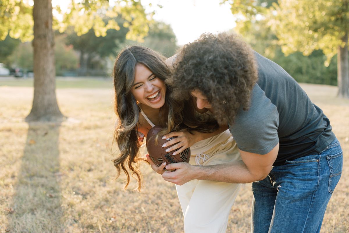 A laughing couple plays with a football outdoors in a sunny park, sharing a joyful and playful moment together.