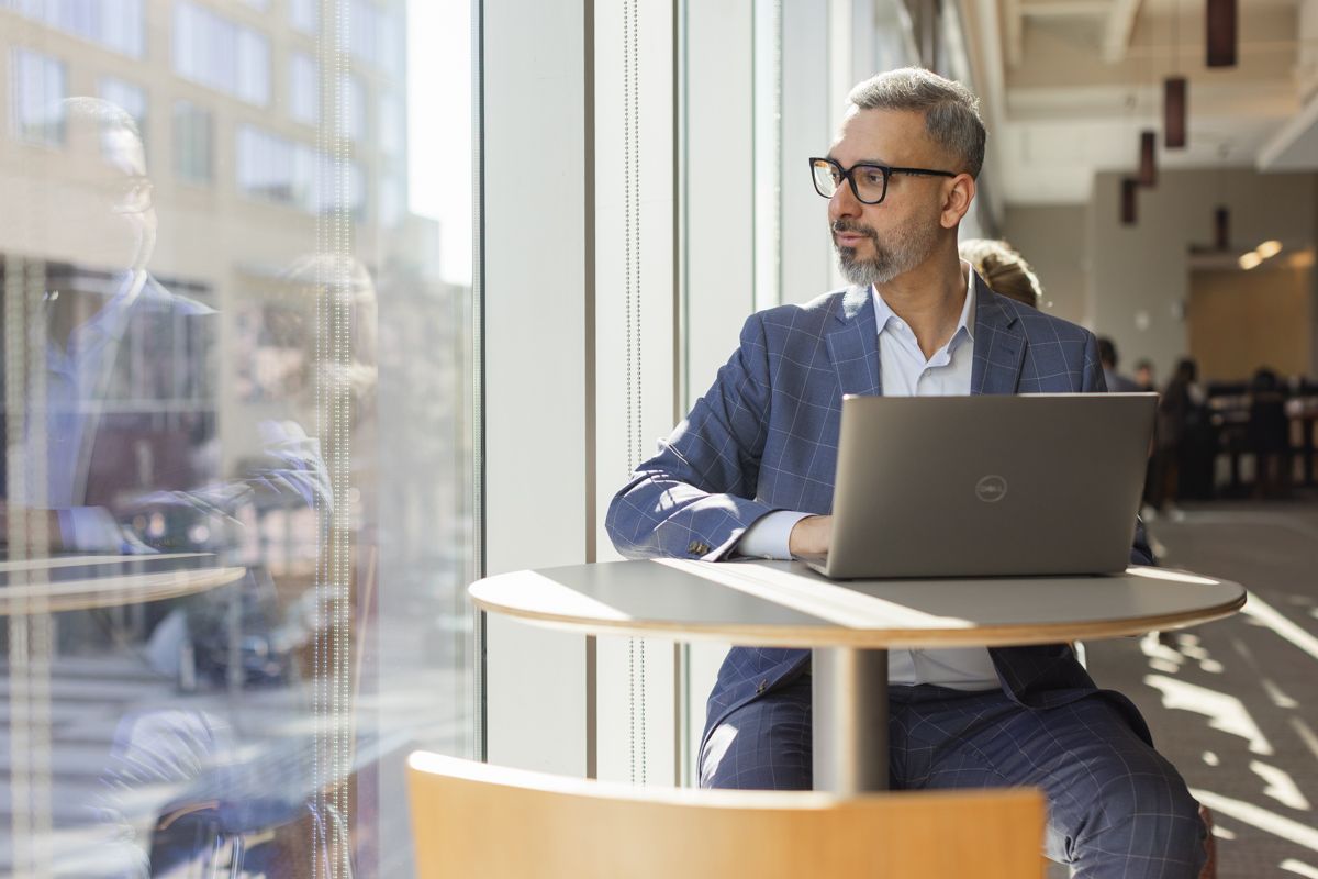 Mature businessman with glasses working on laptop at table by large office windows.