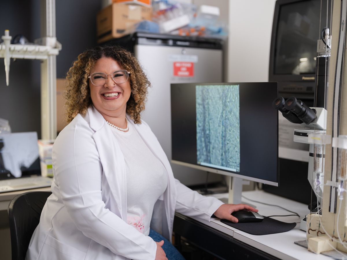 Smiling female scientist in white lab coat sitting at computer with microscope in laboratory.