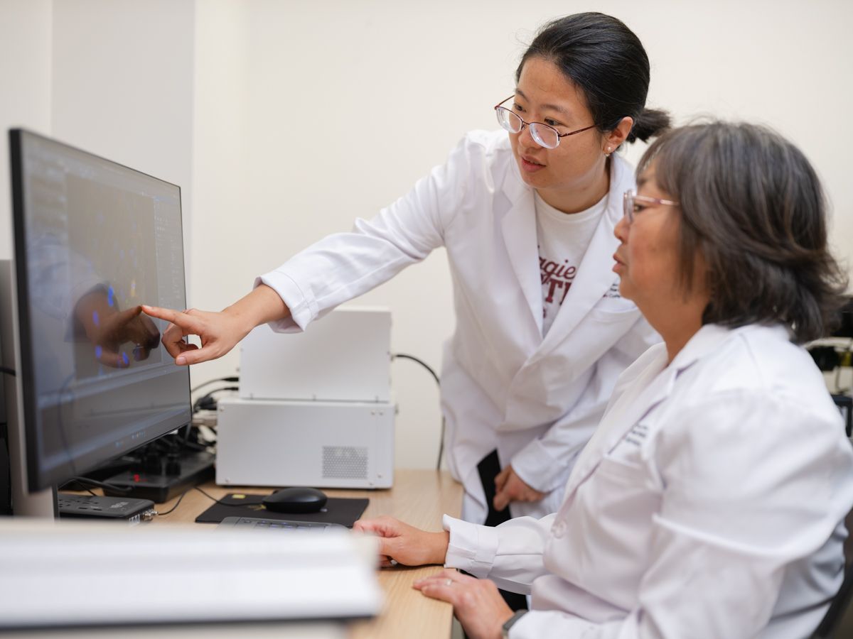 Two female researchers in white lab coats reviewing medical imaging results on computer monitor.