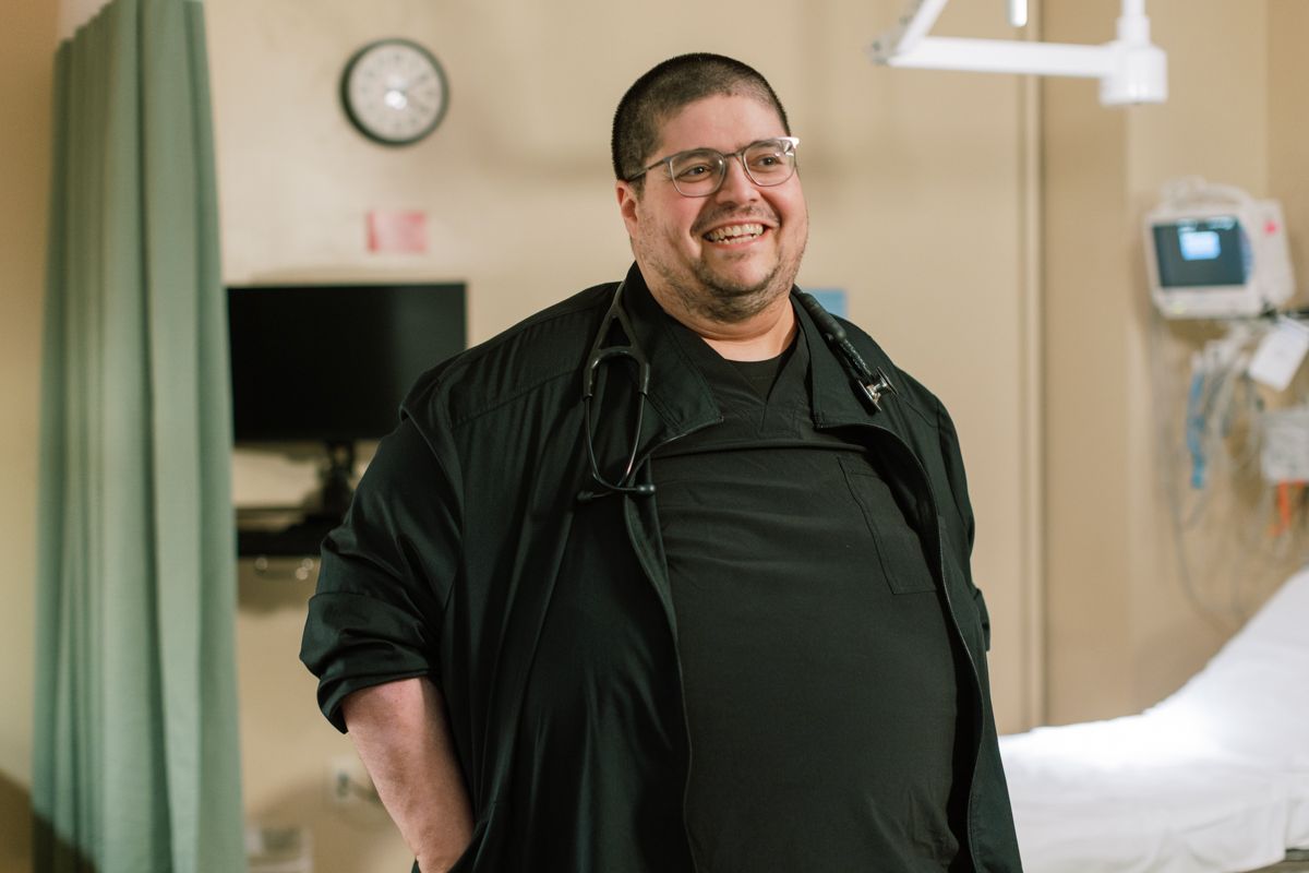 Smiling male healthcare professional with stethoscope standing in hospital room wearing black scrubs and glasses.