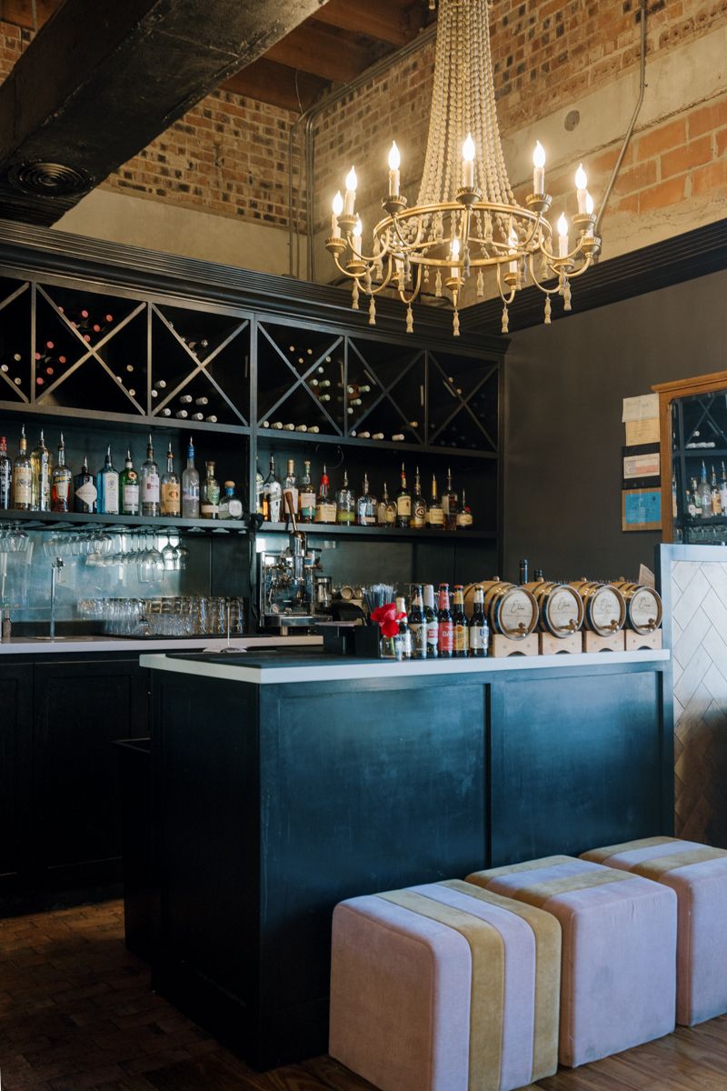 Elegant bar interior with dark cabinetry, gold chandelier, wine rack, and striped ottoman stools on hardwood floors.