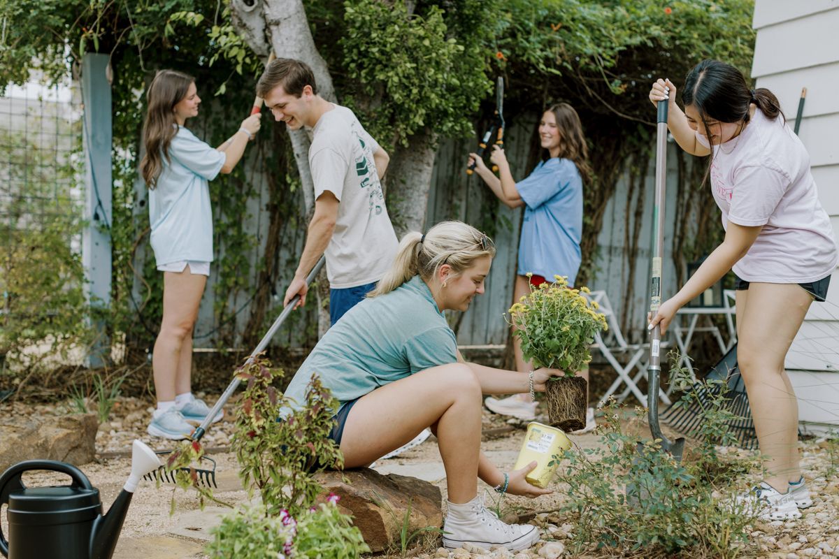 Group of five young volunteers working together raking and gardening in an outdoor yard.