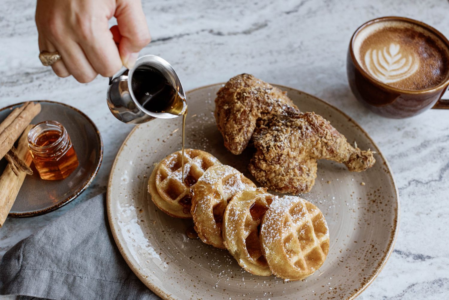 Chicken and waffles with powdered sugar on a ceramic plate, syrup being poured, with a latte and honey on a marble table.
