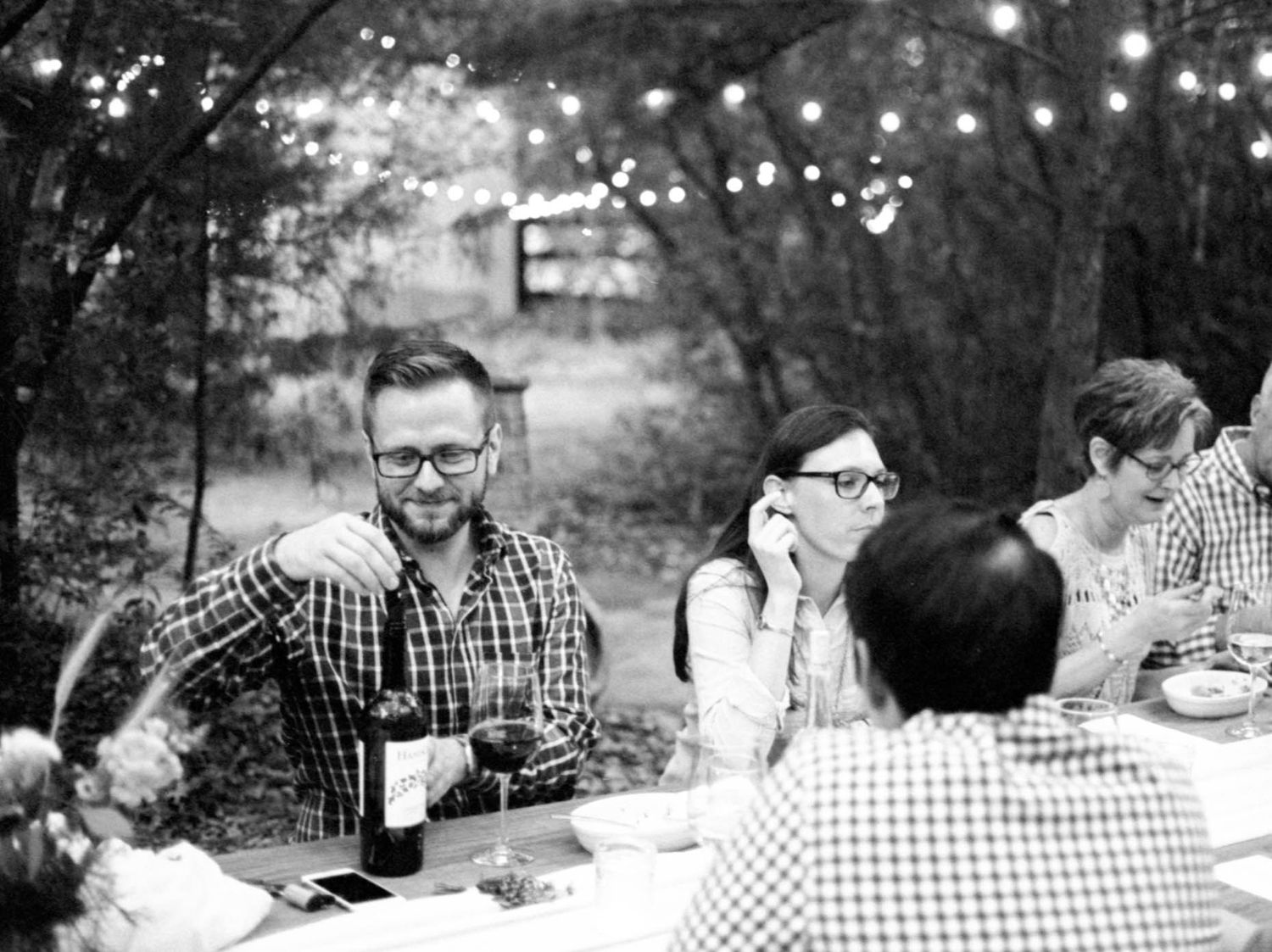 Black and white photo of guests enjoying wine and dinner at an outdoor table with string lights.