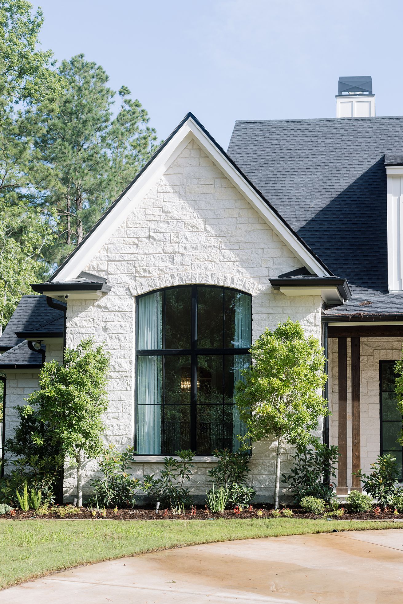 White stone home exterior with arched steel-frame window, black roof, and manicured front yard landscaping.