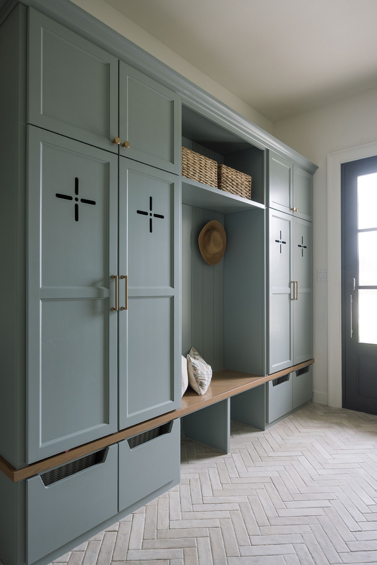 Sage green mudroom with locker-style cabinetry, brass hardware, wood bench, herringbone tile floor, and woven baskets.