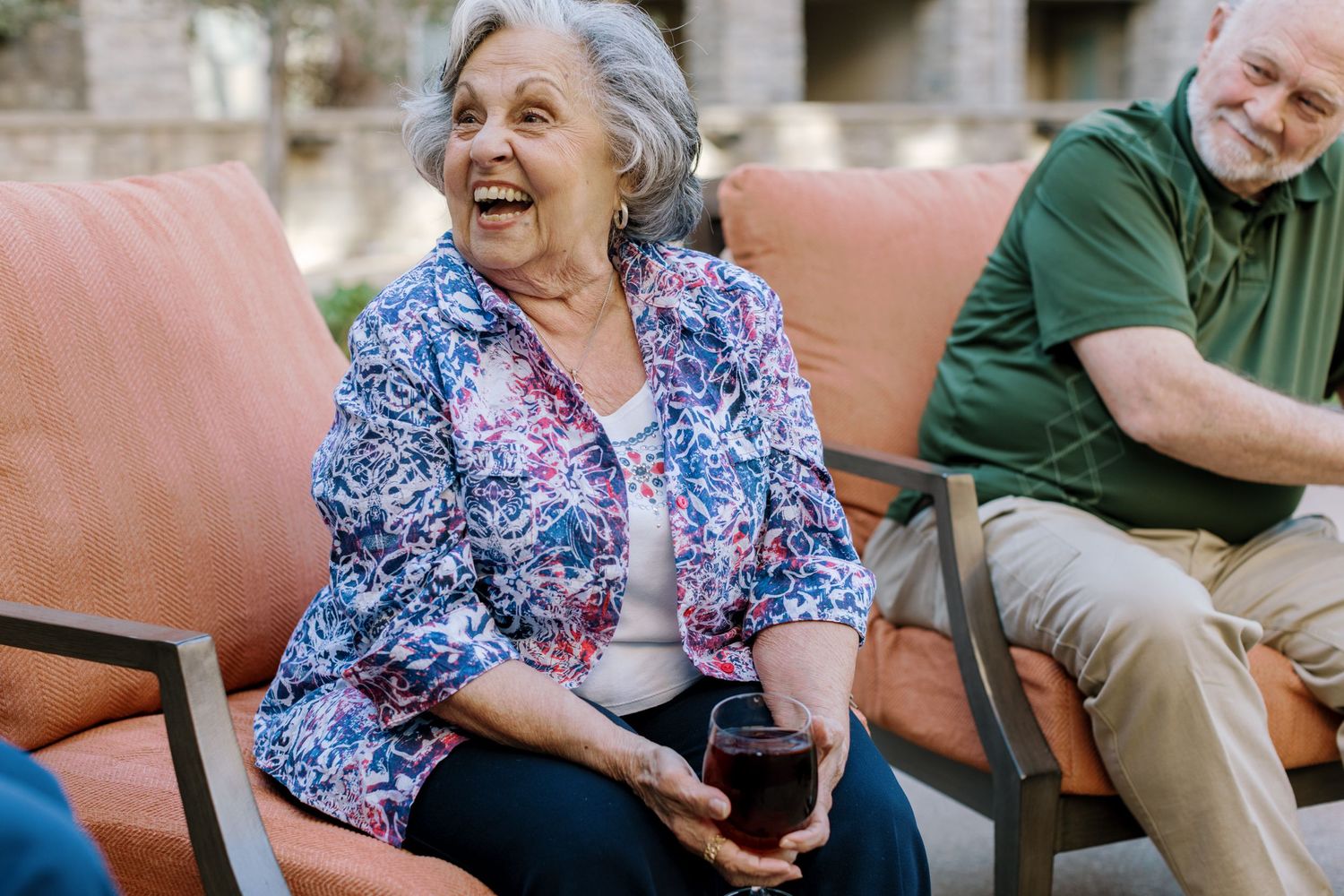 Cheerful senior woman laughing while socializing with friends outdoors on patio chairs.