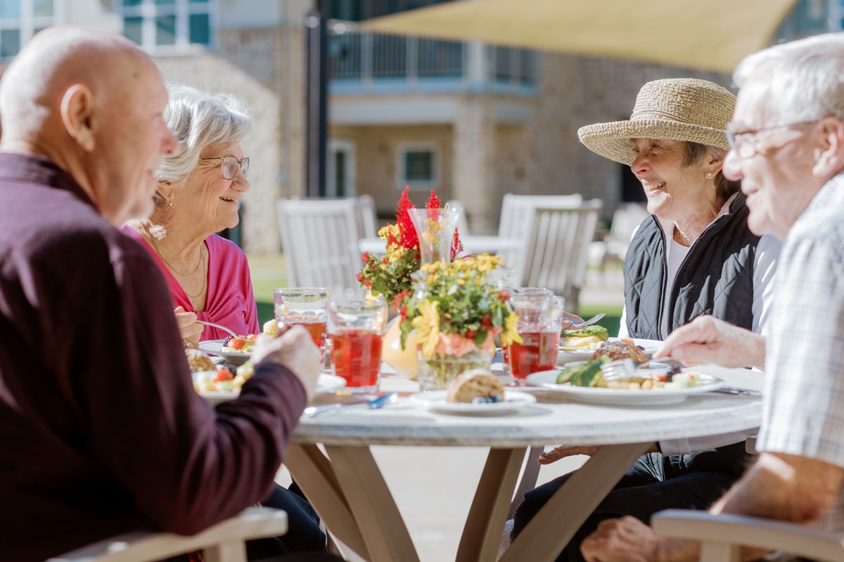 Four seniors enjoying an outdoor meal together at a patio table with flowers and drinks on a sunny day.