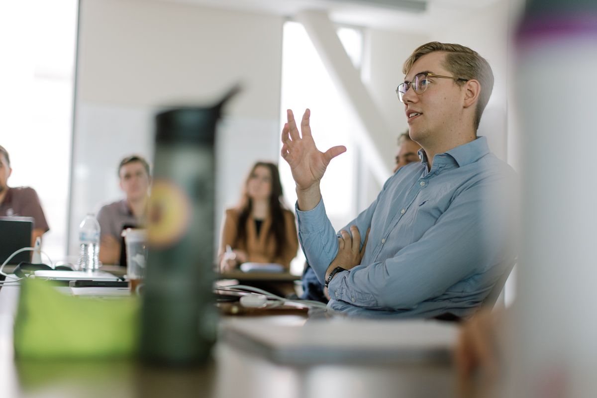 Man in blue shirt gestures while speaking at a business meeting with colleagues in background.