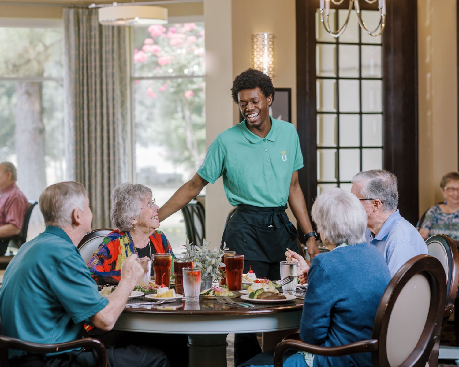 Cheerful server in green polo serving food to group of seniors at elegant dining table.