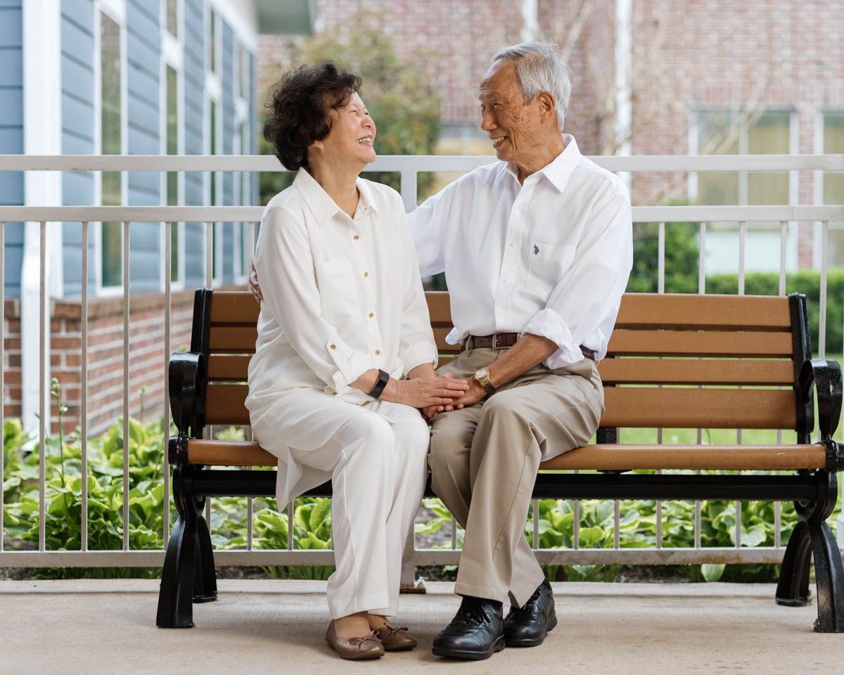 Happy Asian senior couple sitting on an outdoor bench, holding hands and smiling at each other.