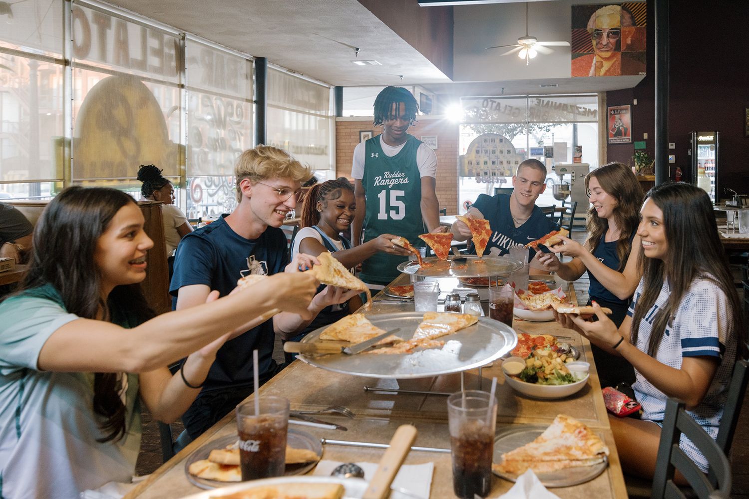 Group of college students in sports jerseys sharing pizza together at a casual restaurant gathering.