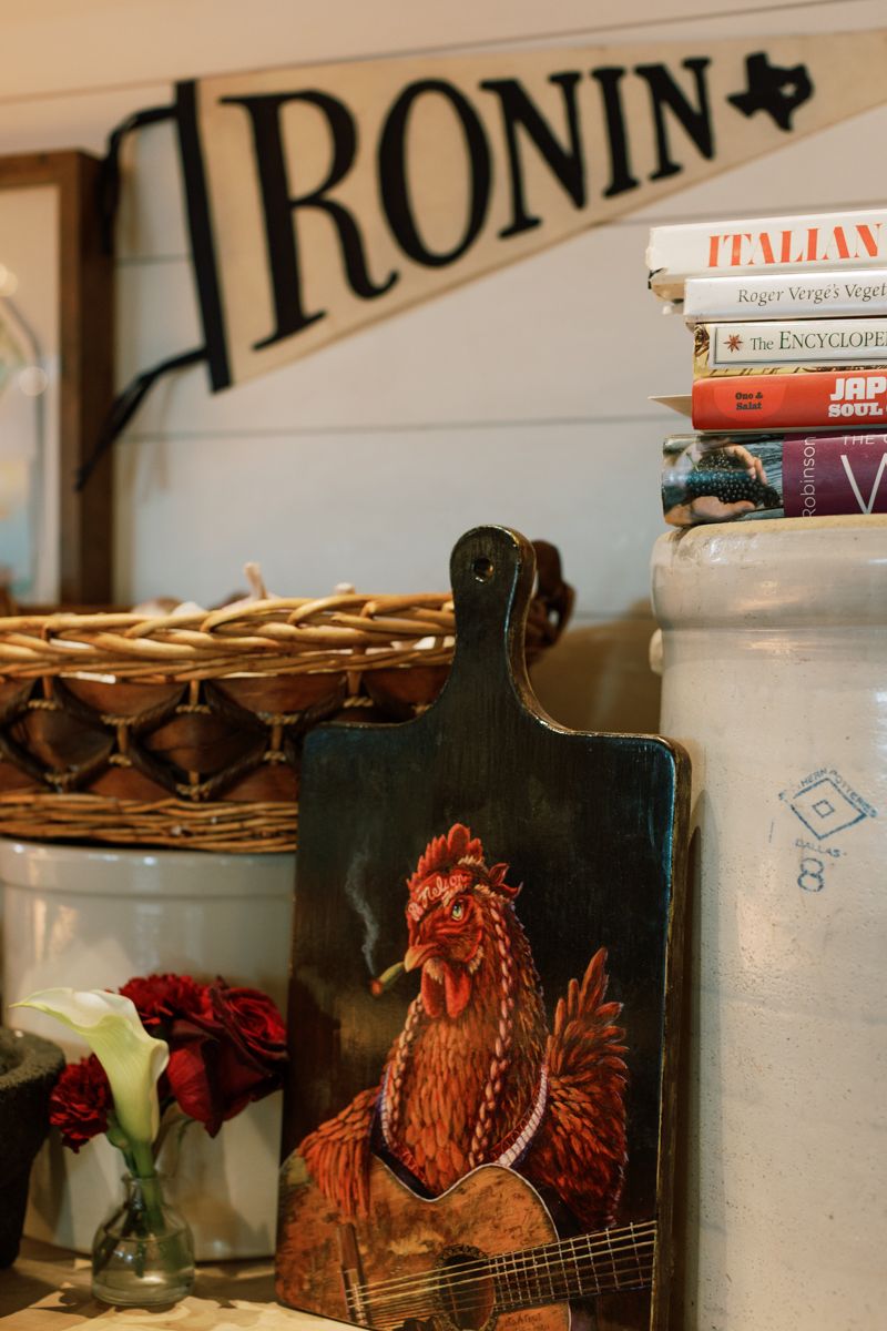 Rustic kitchen shelf with rooster painting on cutting board, cookbooks, wicker basket, and Ronin pennant sign.