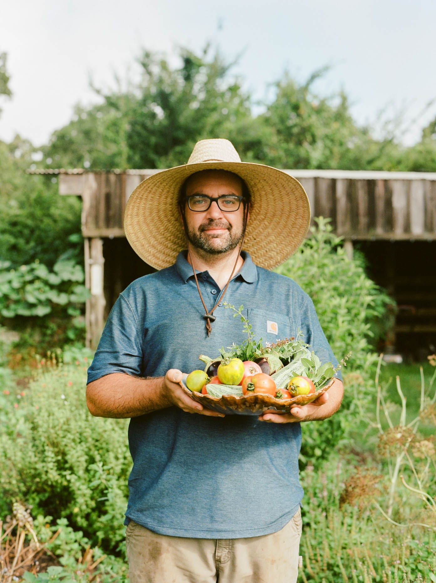 Man in straw hat holding a bowl of fresh garden vegetables outdoors.