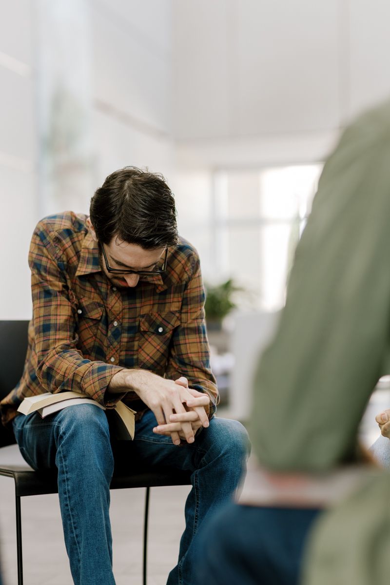 Man in plaid shirt sitting with head bowed, hands clasped over a book in a bright room.