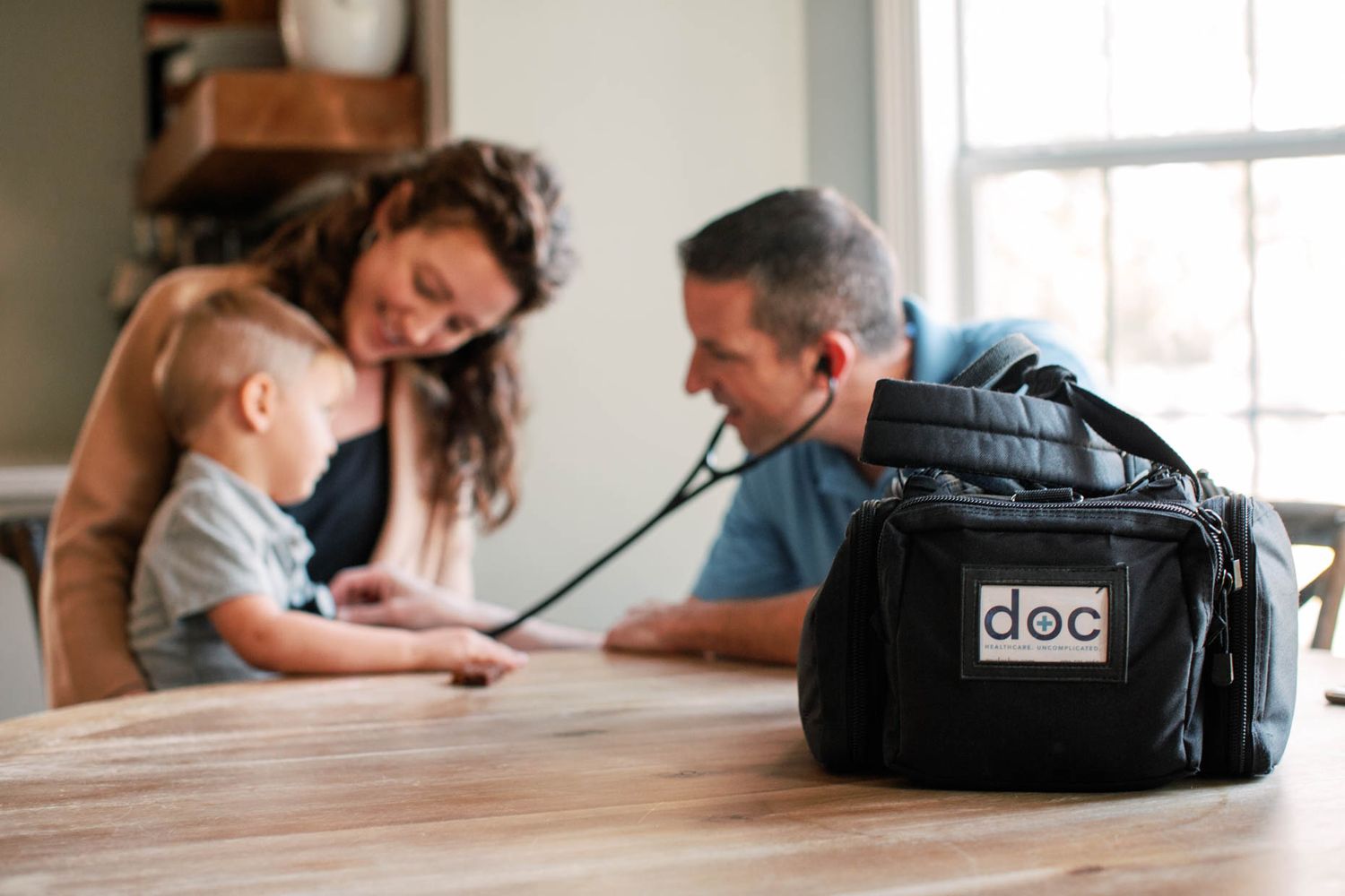 Doctor using stethoscope on baby while parents sit at wooden dining table with medical bag in foreground.