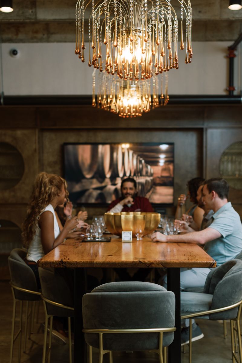 Four people enjoy drinks at a wooden table beneath a gold chandelier in an upscale restaurant.