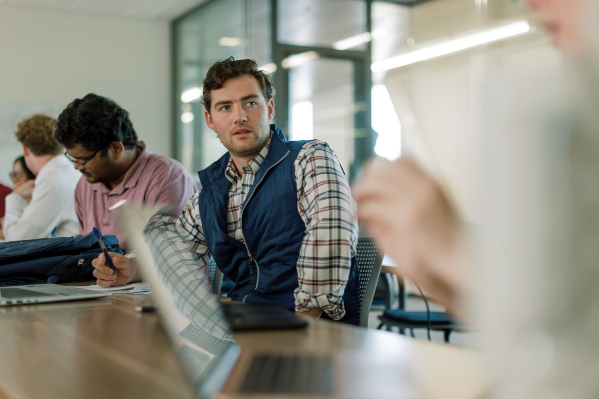 Young man in plaid shirt and blue vest sitting at conference table, looking over shoulder in modern office.