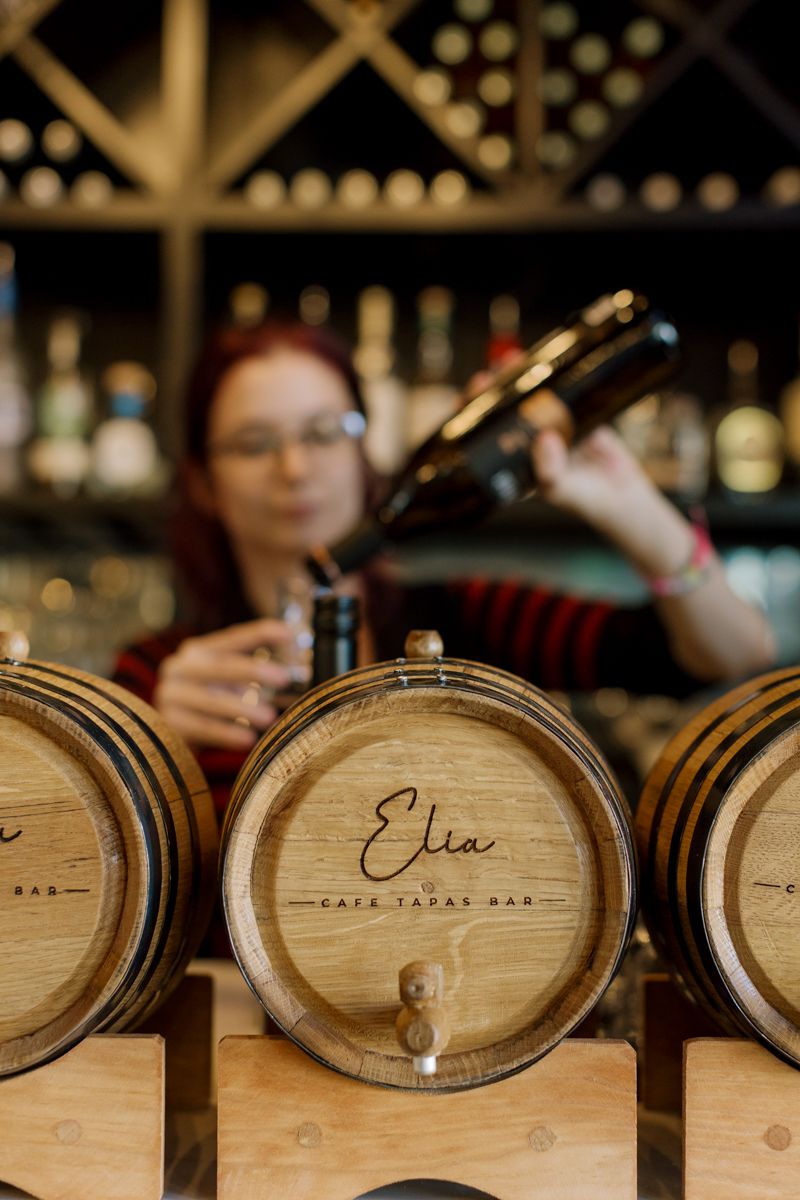 Wooden wine barrels branded with Elia Cafe Tapas Bar logo, with a woman pouring wine in the background.