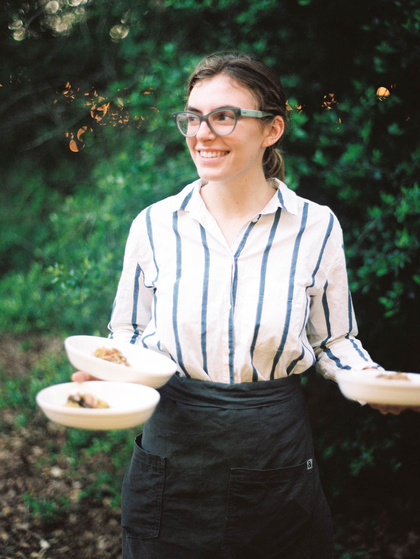 Smiling server in striped shirt and black apron carrying plates of food in a lush garden setting.