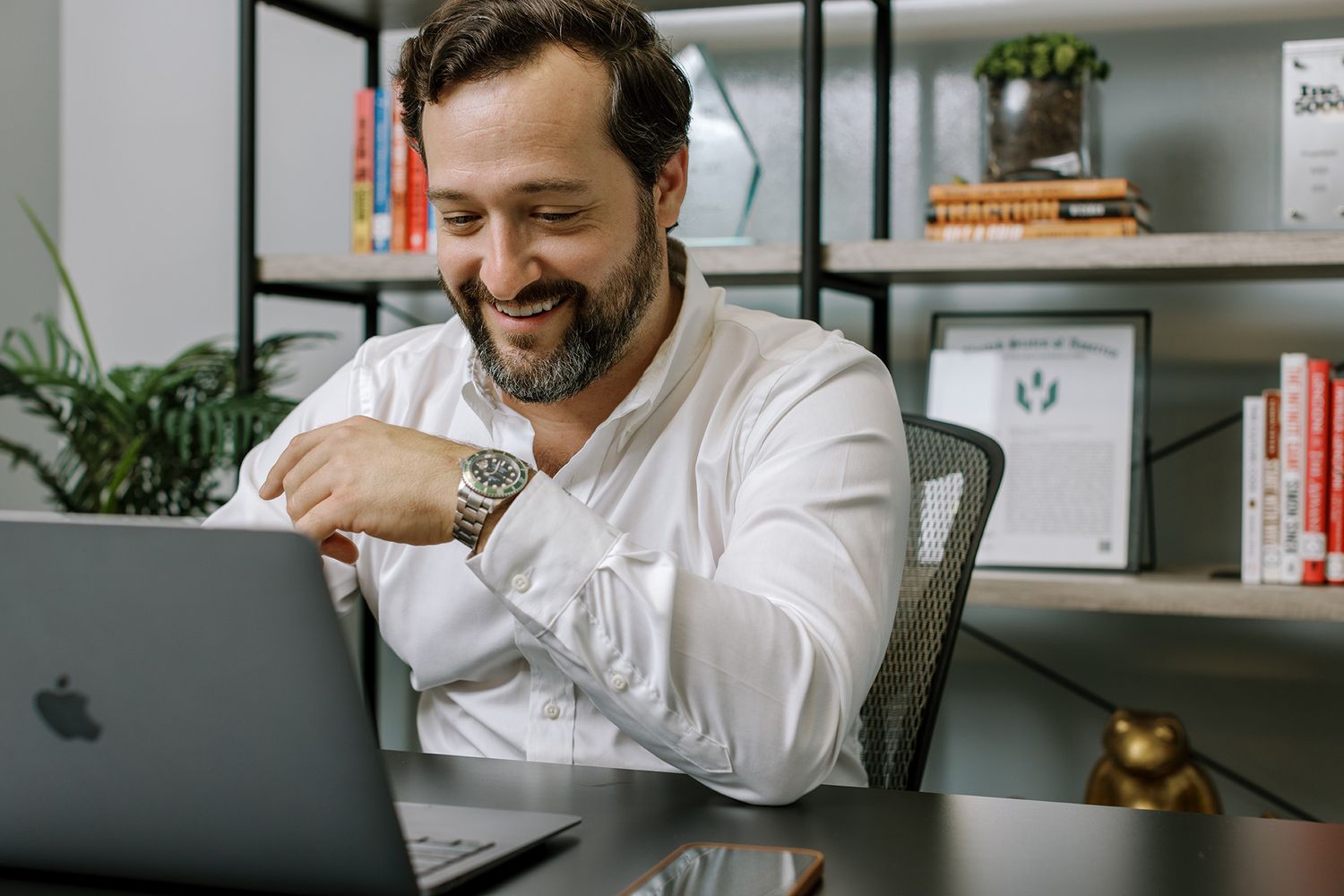 Smiling bearded man in white shirt working on MacBook laptop at desk with bookshelf in modern office.