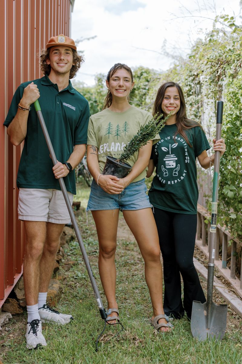 Three students holding gardening tools and a plant while volunteering for a community service planting project outdoors.