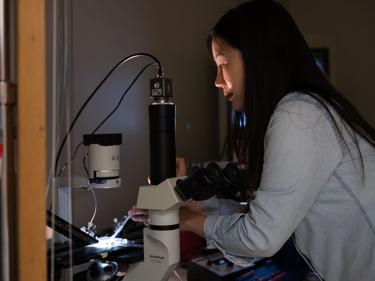 Young female researcher using a microscope in a dimly lit scientific laboratory setting.