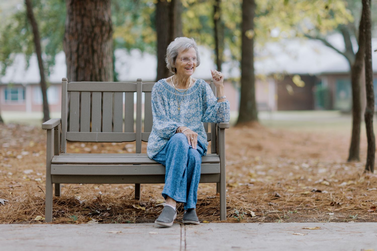 Elderly woman with white hair sitting on a wooden park bench, smiling outdoors among autumn trees.