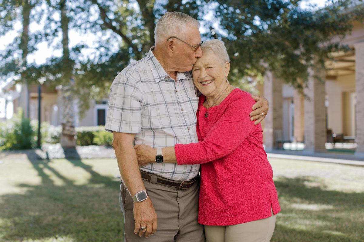 An elderly man kisses his smiling wife on the cheek while embracing outdoors near a building.