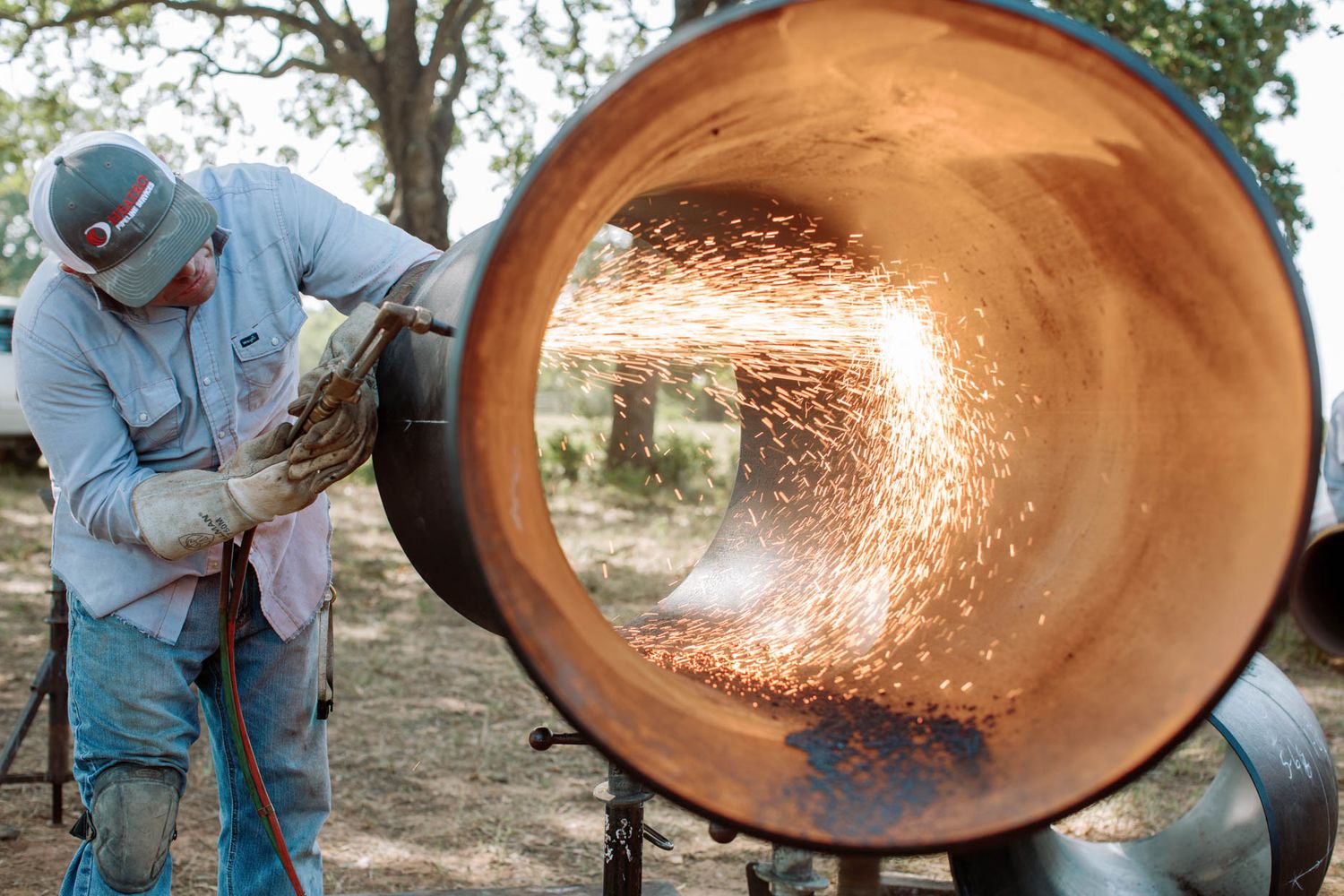 Worker grinding inside large metal pipe with bright sparks flying in outdoor construction setting.