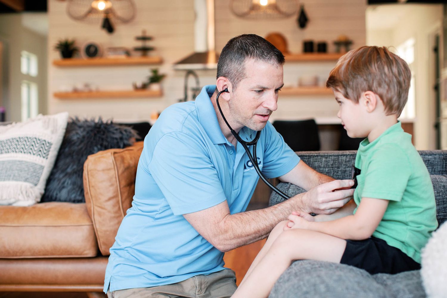 A doctor in a blue polo uses a stethoscope to examine a child on a leather couch at home.