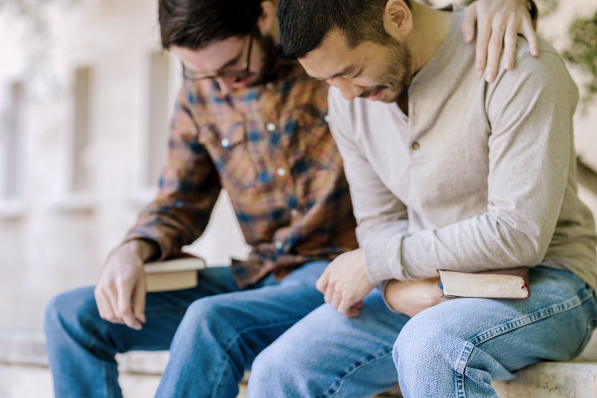 Two men sitting together on steps, one consoling the other with an arm around his shoulder, both holding books.