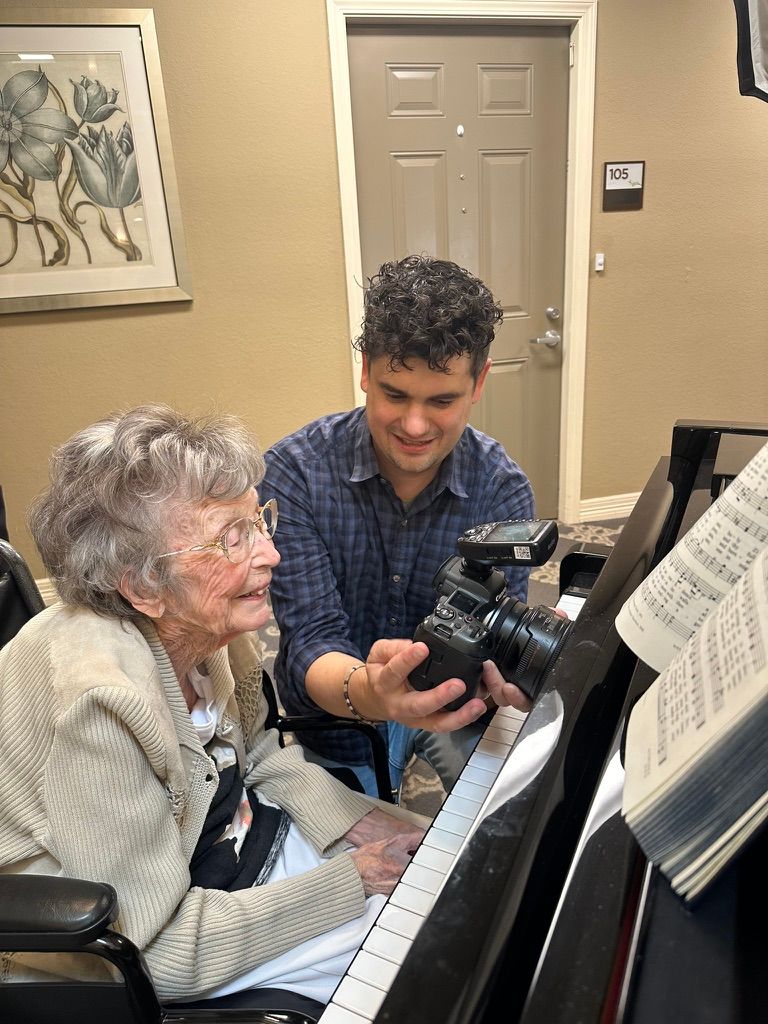 Young photographer shares camera screen with elderly woman seated at a piano indoors.